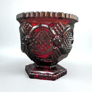 May include: A ruby red glass vase with a detailed, textured pattern. The vase has a square base, a pedestal, and a wide, scalloped rim. The design includes diamond and circular patterns, creating a vintage aesthetic.