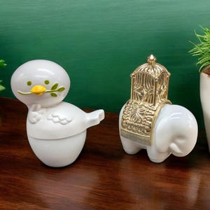 May include: Two white ceramic figurines, one shaped like a bird with a green twig in its beak, the other shaped like an elephant with a gold cage on its back.