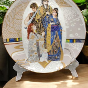 May include: A white porcelain plate with a gold rim features a colorful painting of a family with a king, queen, and two children. The painting is in a style reminiscent of Gustav Klimt's work. The plate is on a decorative stand.