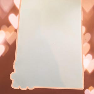 May include: A blank white card with a light peach border. The card is against a background of out-of-focus pink hearts.