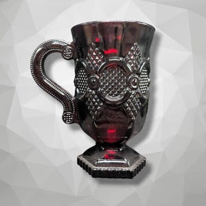 May include: A dark, metallic-looking mug with an ornate handle and decorative detailing. The mug has a pedestal base and a detailed design on the front. The handle is curved and has a decorative element at the top and bottom.