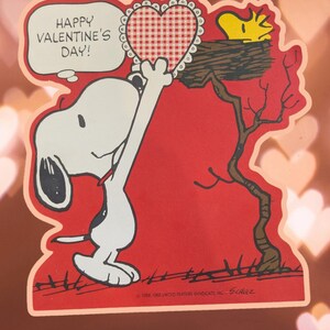 May include: A red Valentine's Day card featuring Snoopy holding a red and white gingham heart. Snoopy is standing on his hind legs and reaching up to give the heart to a yellow bird sitting in a tree branch. The card says "Happy Valentine's Day!"