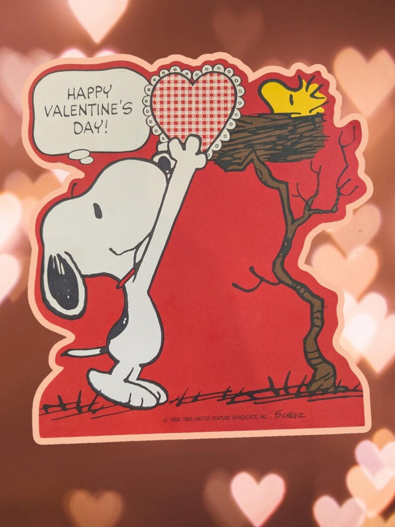 May include: A red Valentine's Day card featuring Snoopy holding a red and white gingham heart. Snoopy is standing on his hind legs and reaching up to give the heart to a yellow bird sitting in a tree branch. The card says "Happy Valentine's Day!"