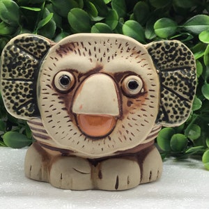 May include: A ceramic koala figurine with a smiling expression. The koala has a beige body with brown accents on the ears, eyes, and stripes. The figurine has a light orange nose and mouth, and is sitting on a base with four feet.