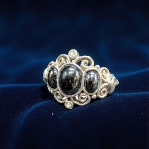 May include: A silver-toned ring featuring three large, oval, black gemstones. The ring has an ornate, filigree design with smaller, clear accent stones. The ring is set against a dark blue background.