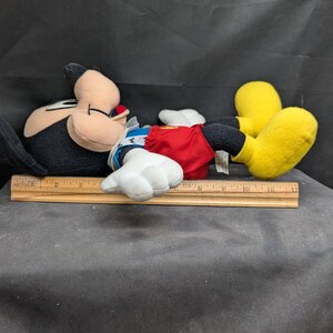 Pu&ograve; includere: Un giocattolo di peluche di Topolino sdraiato su un lato. Topolino indossa un costume rosso e bianco con un colletto blu. Ha scarpe nere con suole gialle.