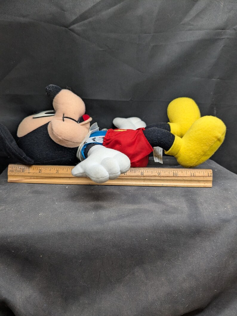 Pu&ograve; includere: Un giocattolo di peluche di Topolino sdraiato su un lato. Topolino indossa un costume rosso e bianco con un colletto blu. Ha scarpe nere con suole gialle.