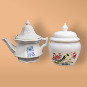 May include: A white teapot with a blue floral design and a white lid. Next to it is a white jar with a lid and a floral design featuring a yellow bird.