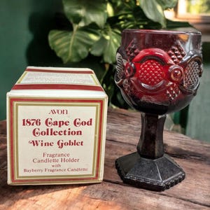 May include: A red glass wine goblet with a cut glass design. The goblet is packaged in a box that reads "Avon 1876 Cape Cod Collection Wine Goblet Fragrance Candle Holder with Bayberry Fragrance Candlelette."
