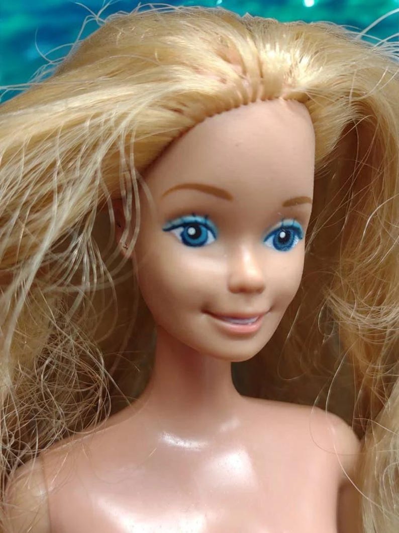 Peut inclure: Gros plan sur le visage d'une poup&eacute;e blonde aux yeux bleus et au sourire. La poup&eacute;e a un teint rose clair et ne porte aucun v&ecirc;tement.