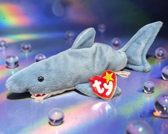 Peut inclure: Une peluche requin bleu clair avec une étiquette rouge en forme de cœur. Le requin a un ventre blanc, un aileron dorsal et une nageoire caudale. L'étiquette indique "ty" en lettres blanches. Le requin est entouré de billes de verre transparentes.