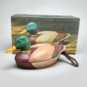 May include: A decorative duck figurine with a green head, yellow beak, and brown body. The duck has a detailed wing design with shades of brown, green, and tan. A brown rope is attached. A box with a duck image is in the background.