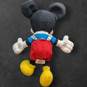 Pu&ograve; includere: Un giocattolo di peluche di Topolino con una camicia blu, una salopette rossa e scarpe gialle. Il giocattolo &egrave; rivolto con le spalle alla telecamera.