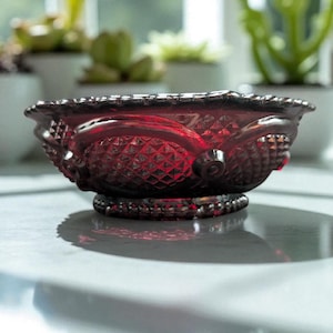May include: A ruby red glass bowl with a decorative, textured design. The bowl features a diamond pattern on the sides and a beaded rim. The bowl is sitting on a white surface.