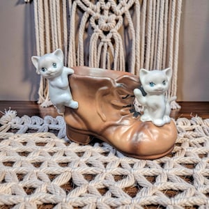 May include: A brown ceramic boot planter with two white cat figurines. One cat is perched on the side, and the other is sitting on the boot. The cats have blue eyes and are in a playful pose. The boot has a worn, vintage look.