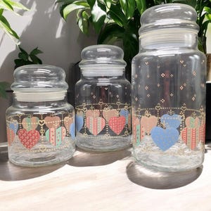 May include: Three glass canisters with lids, featuring a heart pattern in shades of pink, blue, and green. The canisters are decorated with gold accents and a repeating pattern of hearts.