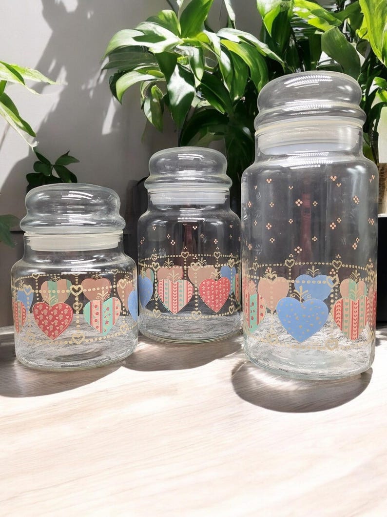 May include: Three glass canisters with lids, featuring a heart pattern in shades of pink, blue, and green. The canisters are decorated with gold accents and a repeating pattern of hearts.