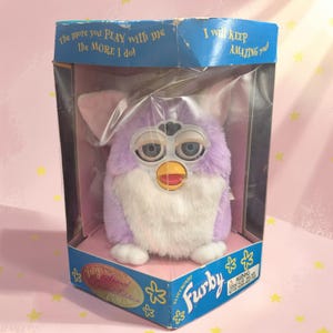 May include: A lavender and white electronic Furby toy in its original blue cardboard packaging. The box features the text The more you PLAY with me the MORE I do! and I will KEEP AMAZING you! The toy has large blue eyes and a yellow beak.