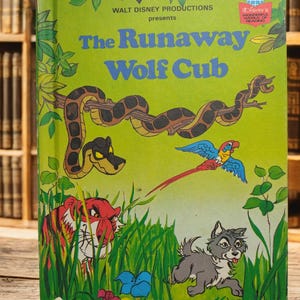 May include: A green book cover with the title "The Runaway Wolf Cub" in blue and white letters. The book is a Walt Disney Productions presentation and features a cartoon wolf cub, a tiger, a snake, and a parrot.