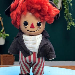 May include: A vintage rag doll with red yarn hair, a black hat, a black jacket, and red and white striped pants. The doll has a smiling face with black eyes and a red nose.