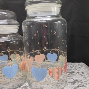 May include: Two clear glass jars with lids, decorated with a pattern of hearts in pink, blue, and green. The jars have a white background with a gold trim.