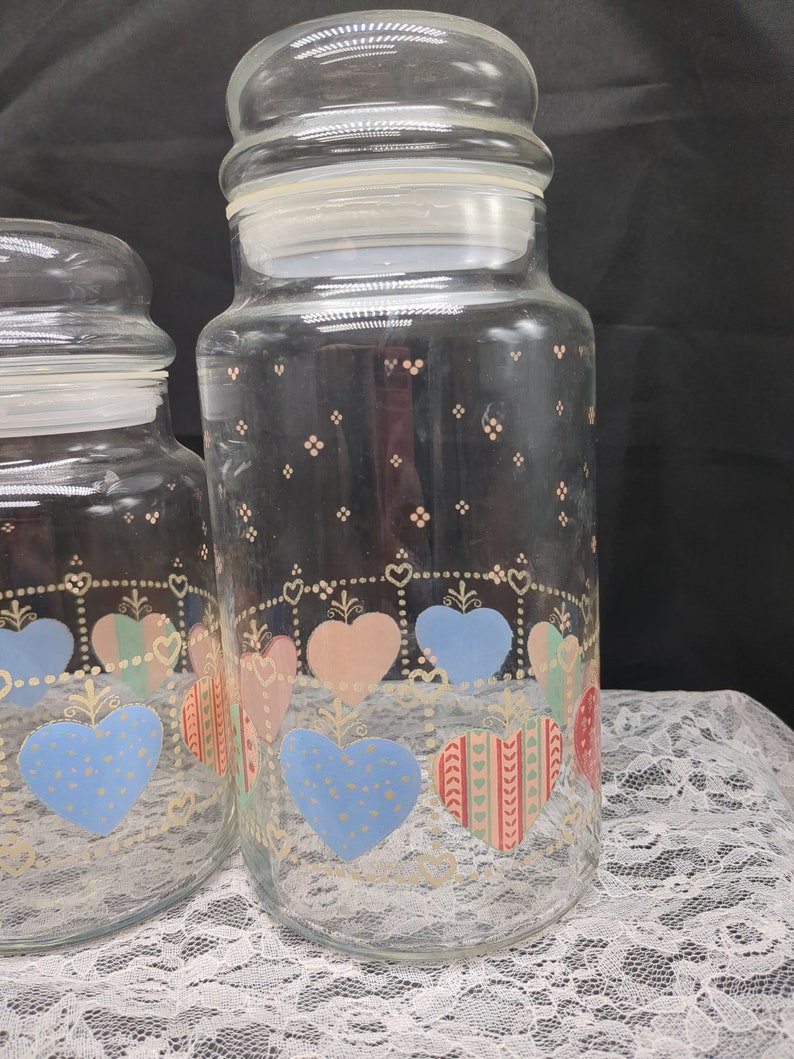 May include: Two clear glass jars with lids, decorated with a pattern of hearts in pink, blue, and green. The jars have a white background with a gold trim.