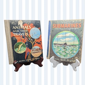 May include: Two vintage children's books on display stands. The book on the left is titled "Animals and Their Travels" with a picture of a fish on the cover. The book on the right is titled "Submarines" with a picture of a submarine in a periscope view.