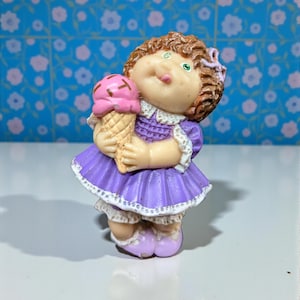 May include: A vintage figurine of a doll in a purple dress, holding a pink ice cream cone. The doll has brown curly hair, green eyes, and a light skin tone. The figurine is a collectible item.