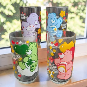 May include: Four clear glass tumblers featuring Care Bears characters. One glass shows Share Bear, one shows Bedtime Bear, one shows Good Luck Bear, and one shows Cheer Bear. Each glass is decorated with colorful hearts, stars, and character names.