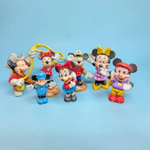 Op de afbeelding: Een verzameling van zeven Mickey en Minnie Mouse beeldjes in verschillende outfits. De beeldjes zijn felgekleurd, met Mickey in een cowboyhoed en Minnie in een rode jurk. Andere personages zijn gekleed in matrozen- en sportkleding.