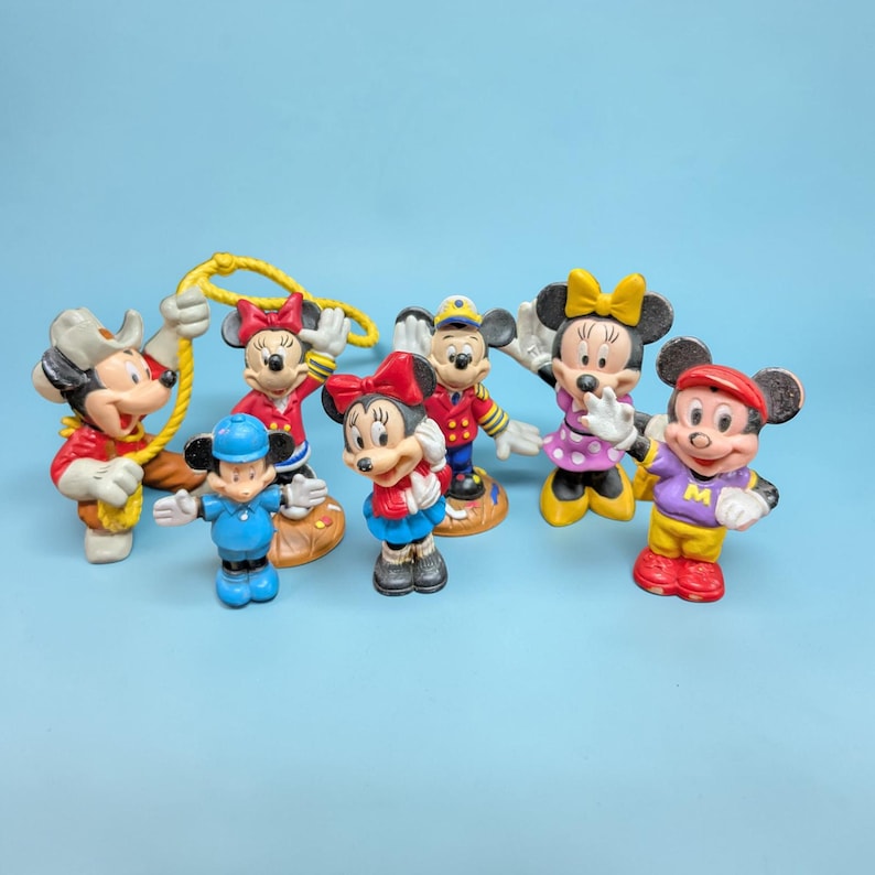 Op de afbeelding: Een verzameling van zeven Mickey en Minnie Mouse beeldjes in verschillende outfits. De beeldjes zijn felgekleurd, met Mickey in een cowboyhoed en Minnie in een rode jurk. Andere personages zijn gekleed in matrozen- en sportkleding.