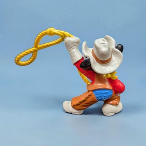 Op de afbeelding: Een beeldje van een cowboy-personage, met een witte hoed, rood overhemd, bruine broek en witte handschoenen, die een geel touw vasthoudt. Op een blauwe achtergrond.