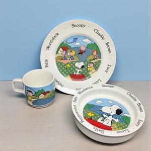 May include: A three-piece children's dish set featuring Snoopy and the Peanuts gang. The set includes a plate, a bowl, and a mug, all with colorful illustrations of the characters. The plate measures approximately 8 inches in diameter.