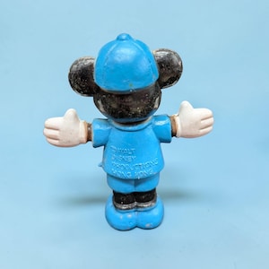 Op de afbeelding: Een vintage Mickey Mouse beeldje in een blauw uniform en pet, met witte handschoenen en uitgestrekte armen. Het beeldje heeft zwarte oren en een blauwe basis. De tekst "WALT DISNEY PRODUCTIONS HONG KONG" is zichtbaar.