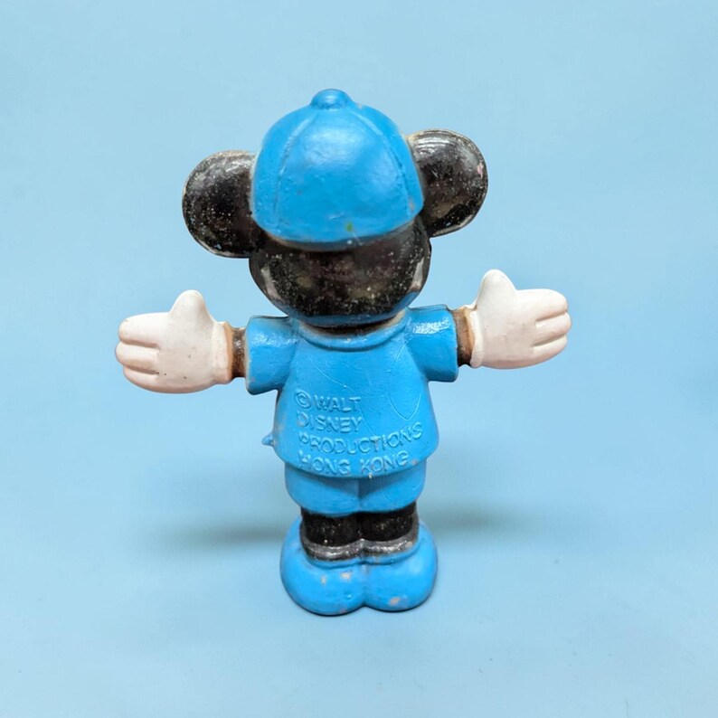 Op de afbeelding: Een vintage Mickey Mouse beeldje in een blauw uniform en pet, met witte handschoenen en uitgestrekte armen. Het beeldje heeft zwarte oren en een blauwe basis. De tekst "WALT DISNEY PRODUCTIONS HONG KONG" is zichtbaar.