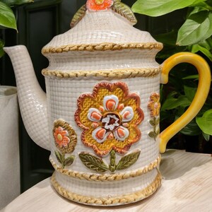 May include: A white ceramic teapot with a floral design. The teapot has a yellow, orange, and red flower design with green leaves. The teapot has a woven rope design around the top and bottom.