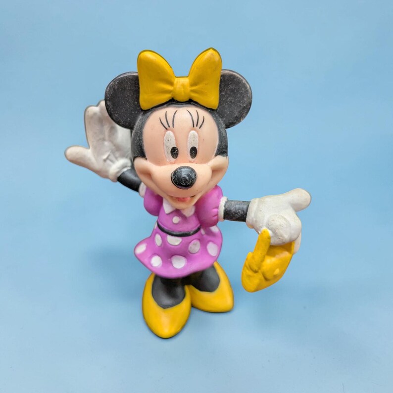 Op de afbeelding: Een Minnie Mouse-beeldje in een zwaaiende pose. De figuur heeft een gele strik, zwarte oren en een roze jurk met witte stippen. Haar schoenen zijn geel en haar handschoenen zijn wit. De achtergrond is effen lichtblauw.