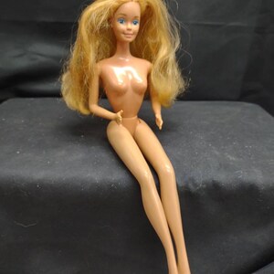Peut inclure: Une poup&eacute;e en plastique nue avec de longs cheveux blonds et des yeux bleus. La poup&eacute;e est assise les jambes crois&eacute;es et les bras reposant sur ses genoux.