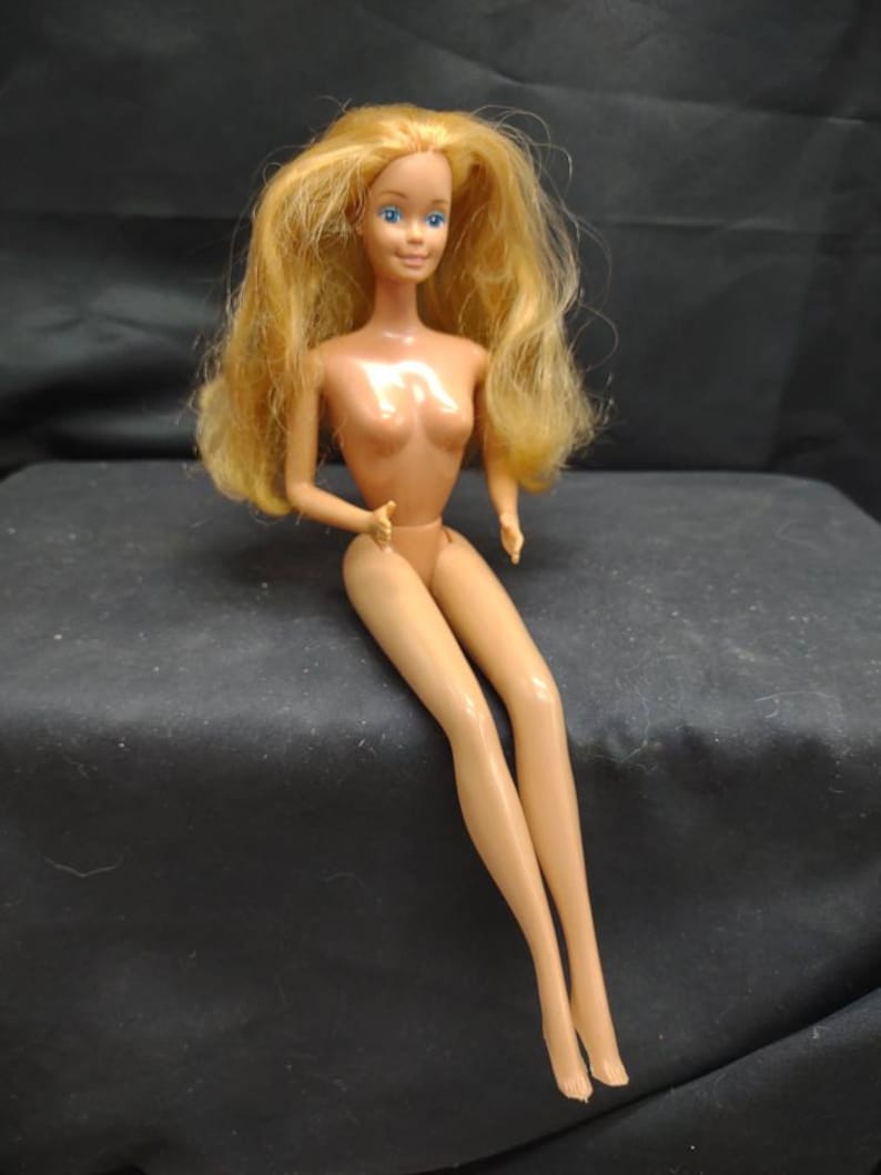 Peut inclure: Une poup&eacute;e en plastique nue avec de longs cheveux blonds et des yeux bleus. La poup&eacute;e est assise les jambes crois&eacute;es et les bras reposant sur ses genoux.