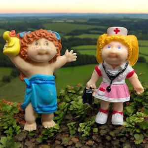 May include: Two Cabbage Patch Kids dolls, one wearing a blue towel and holding a yellow rubber ducky, the other wearing a pink and white nurse's uniform and holding a black medical bag.