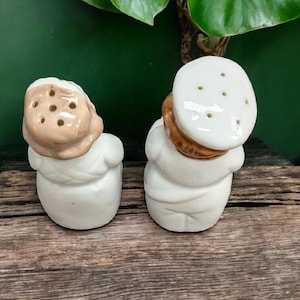 May include: A pair of ceramic salt and pepper shakers shaped like people. The shakers are white with brown accents and have holes on top for dispensing salt and pepper.