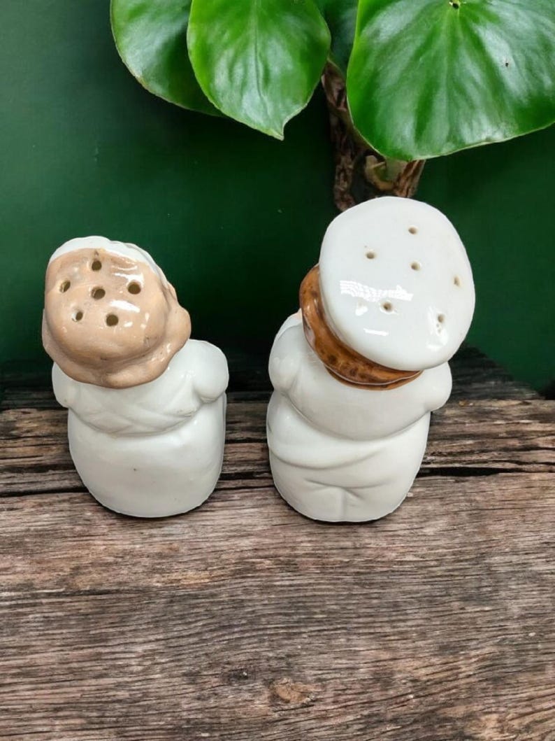 May include: A pair of ceramic salt and pepper shakers shaped like people. The shakers are white with brown accents and have holes on top for dispensing salt and pepper.