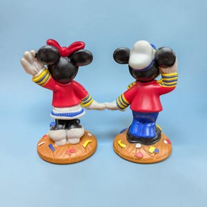 Op de afbeelding: Twee beeldjes van Mickey en Minnie Mouse die elkaars hand vasthouden. Minnie draagt een rode jurk, een rode strik en zwarte schoenen. Mickey draagt een rode jas, een blauwe broek en een witte matrozenhoed. Beide staan op bruine voetstukken met confetti.