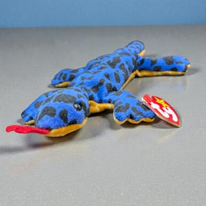 May include: A blue plush lizard toy with black spots and a red tongue. The toy has yellow accents and a red tag attached. The lizard is lying on a gray surface.