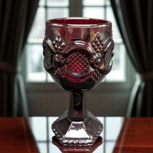 May include: A ruby-red, vintage-style goblet with ornate detailing. The glass features a textured pattern around the bowl and a faceted base. The goblet is displayed on a polished wooden surface, with a window and curtains in the background.