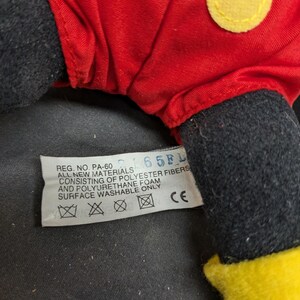 Pu&ograve; includere: Un giocattolo di peluche rosso e nero con un viso giallo. Il giocattolo ha un'etichetta che recita "REG. NO. PA-60 65FL ALL NEW MATERIALS CONSISTING OF POLYESTER FIBERS AND POLYURETHANE FOAM SURFACE WASHABLE ONLY CE".