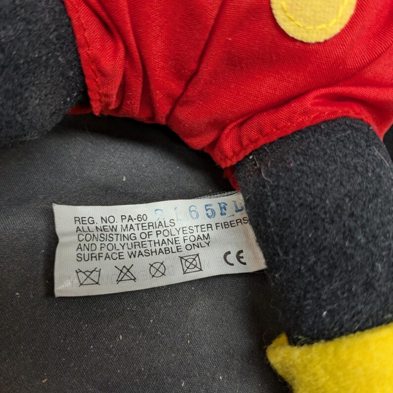 Pu&ograve; includere: Un giocattolo di peluche rosso e nero con un viso giallo. Il giocattolo ha un'etichetta che recita "REG. NO. PA-60 65FL ALL NEW MATERIALS CONSISTING OF POLYESTER FIBERS AND POLYURETHANE FOAM SURFACE WASHABLE ONLY CE".
