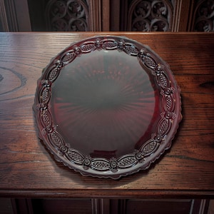 May include: A round, ruby-red glass serving tray with a decorative, embossed border. The tray has a scalloped edge and a central, radiating pattern. The tray is displayed on a dark wooden surface.