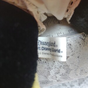 Op de afbeelding: Close-up van een zwart en geel pluche speelgoed met een witte kantversiering. Het speelgoed heeft een wit label met de tekst "Disneyland&reg; Walt Disney World&reg; Walt Disney Productions&reg;".