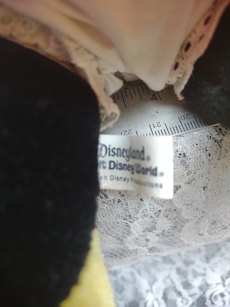 Op de afbeelding: Close-up van een zwart en geel pluche speelgoed met een witte kantversiering. Het speelgoed heeft een wit label met de tekst "Disneyland&reg; Walt Disney World&reg; Walt Disney Productions&reg;".